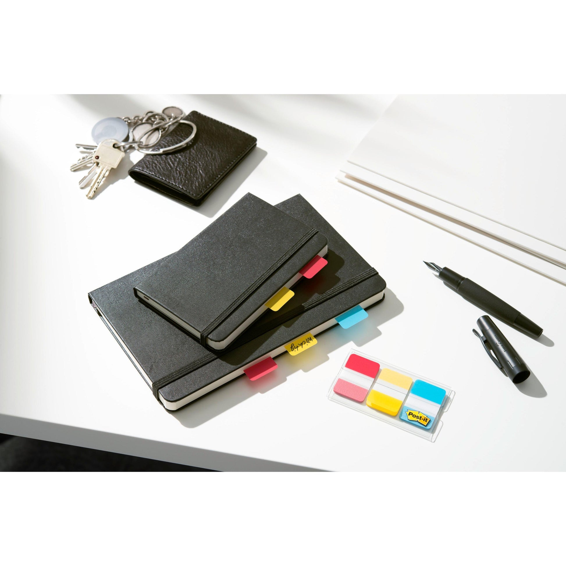 Ein Satz schwarzer Notizbücher mit Post-it® Index Strong Ablagetabs der 3M Deutschland GmbH in verschiedenen Farben, eine Brieftasche, Schlüssel, weißes Papier, ein schwarzer Stift und ein Marker liegen auf einem weißen Schreibtisch.