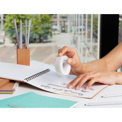 A person uses correction tape over handwritten notes in a spiral notebook, surrounded by office supplies like pencils, pens, and 3M Deutschland GmbH Scotch® Magic™ Tape (66% plant-based adhesive, 19 mm x 33 m) at a window overlooking a city street.