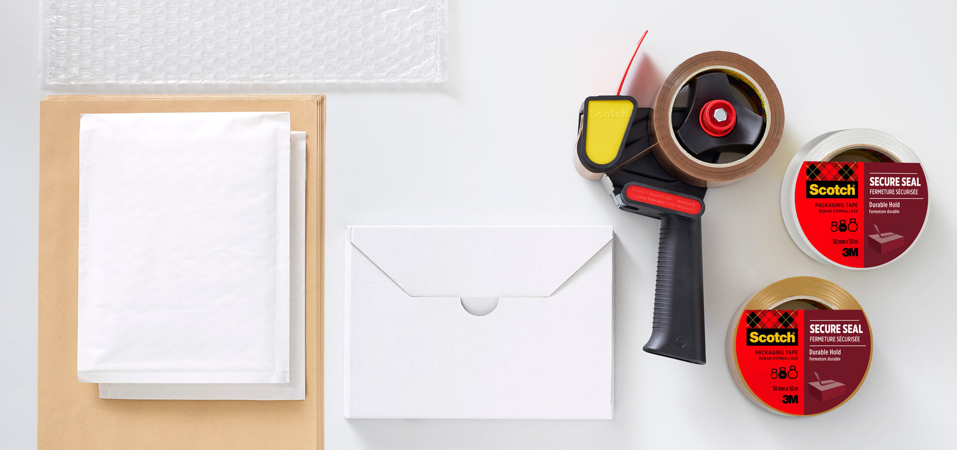 Eine flache Lage Verpackungsmaterial, einschließlich Luftpolsterfolie, Papierbögen, braune Umschläge, eine geschlossene weiße Schachtel, Klebebandabroller und zwei Rollen 3M Deutschland GmbH Scotch® Verpackungsklebeband für einen sicheren Verschluss (50 mm x 50 m).