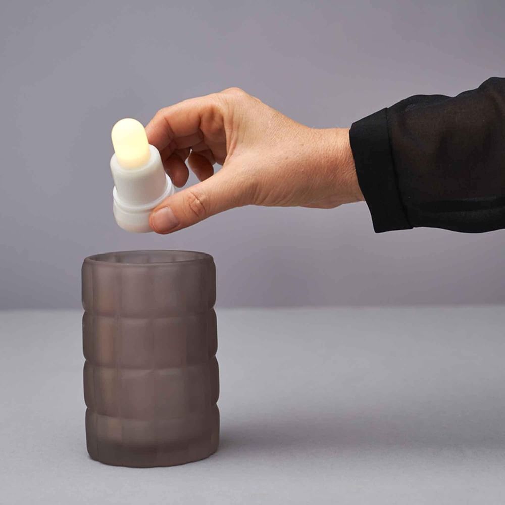 A hand holds a small, rechargeable LED candle from the LED Set Warm White & Multicolor (Duni GmbH, 12 pieces, color changing) with a rounded tip over a dark cup on a gray surface against a plain background.
