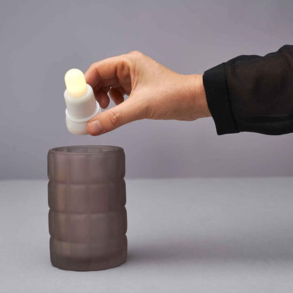 A hand holds a small, rechargeable LED candle from the LED Set Warm White & Multicolor (Duni GmbH, 12 pieces, color changing) with a rounded tip over a dark cup on a gray surface against a plain background.