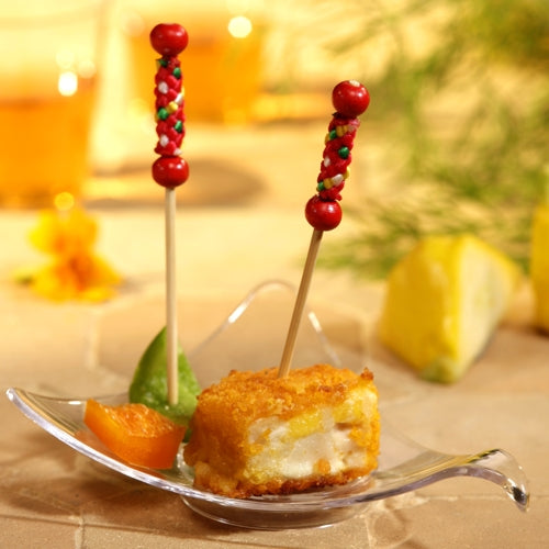 Auf einem transparenten, blattförmigen Teller liegt ein Würfel panierter, frittierter Speisen mit einem PAPSTAR 250 Fingerfood-Picker der PAPSTAR GmbH, dazu eine grüne und eine Orangenscheibe. Vor dem leicht verschwommenen Hintergrund sind eine Zitronenscheibe und Grünzeug zu sehen.