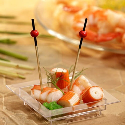 On a transparent, square dish are imitation crab, shrimp, a green garnish, and two PAPSTAR 250 finger food picks from PAPSTAR GmbH with red beads. In the background, blurred shrimp can be seen on a plate.