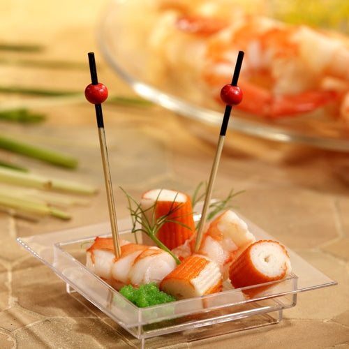 Auf einer durchsichtigen, quadratischen Schale befinden sich Krabbenimitat, Garnelen, eine grüne Garnitur und zwei PAPSTAR 250 Fingerfood-Picker der PAPSTAR GmbH mit roten Perlen. Im Hintergrund sind auf einem Teller verschwommene Garnelen zu sehen.