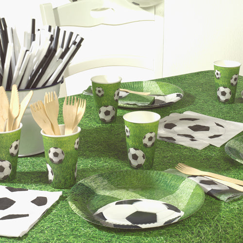 Ein Partytisch wird mit PAPSTAR 3-lagigen Fußballservietten (20 Servietten, 33x33 cm, Design Set 4), schwarz-weiß gestreiften Strohhalmen, Holzbesteck in einem Halter und einer grünen Tischdecke in Grasform gedeckt. Marke: PAPSTAR GmbH.
