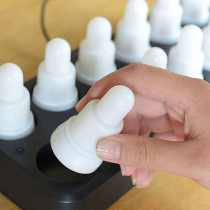 A hand places or removes a white, round LED candle from the black tray of the Duni GmbH LED Set, Warm White & Multicolor - 12 Piece Color Changing Lights; each identical, rechargeable candle sits on a wooden base.