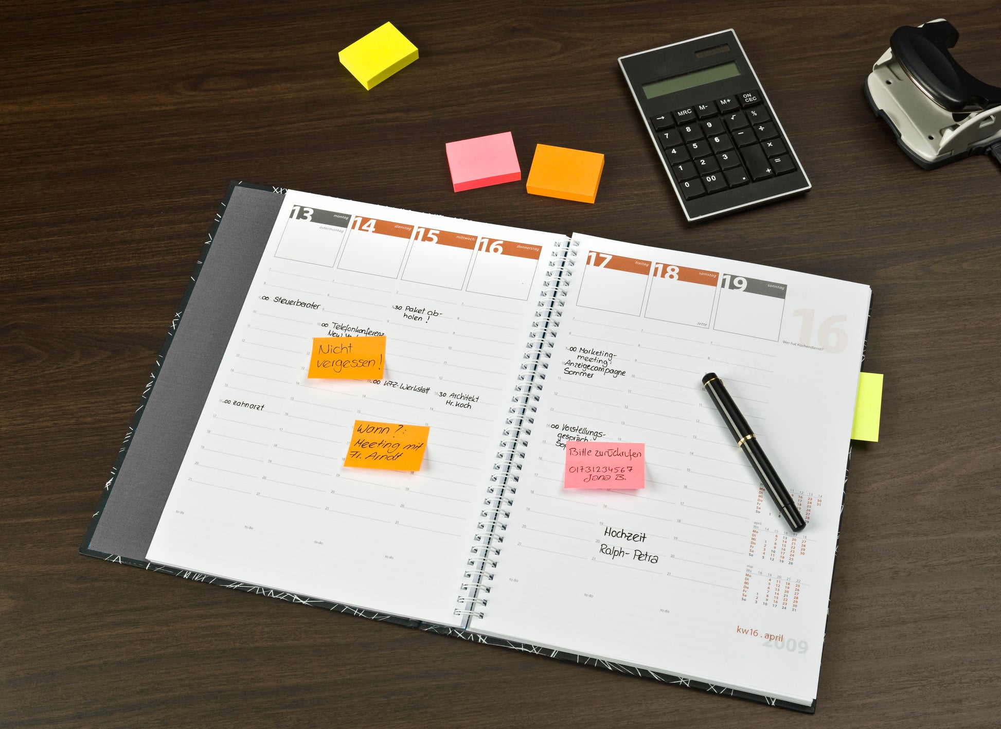Ein Wochenplaner liegt offen auf einem Schreibtisch, an dem bunte 3M Deutschland GmbH Post-it® Zettel in verschiedenen Neonfarben (38 mm x 51 mm) befestigt sind. Ein Stift liegt darauf, daneben ein Taschenrechner, ein Hefter und zwei Stapel neonfarbener Haftnotizen.