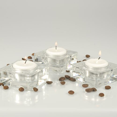 Drei Starpak Teelichter Ø 38 mm, weiß, mit 8 h Brenndauer aus der Faltschachtel (100 Stück) stehen in quadratischen Glasbehältern auf weißer Fläche mit verstreuten Kaffeebohnen vor hellem, schlichten Hintergrund.