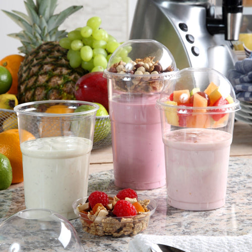 Drei Smoothie-Becher stehen auf einer Theke, zwei mit Obst und Granola in Domdeckeln. Die Deckel bestehen aus Starpak 50 Einsätzen, PET „Hurricane“ rund Ø 9 cm, glasklar. Im Hintergrund stehen frische Ananas, Trauben und ein Mixer.