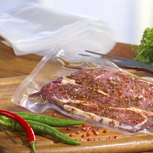 Ein Starpak 100 Siegelrandbeutel, PA/PE transparent von Starpak mit mariniertem rohem Fleisch liegt auf einem hölzernen Schneidebrett, umgeben von roten und grünen Chilischoten und Blattgemüse im Hintergrund.