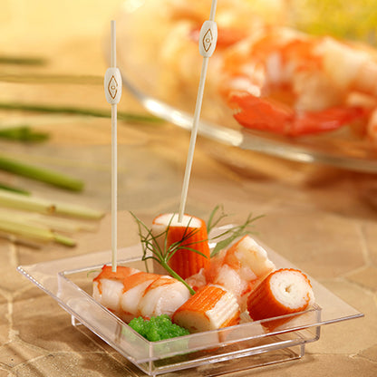 On a square plastic tray with imitation crab, shrimp, green roe, and garnish, two PAPSTAR 40 finger food picks "pure" from PAPSTAR GmbH are depicted, with a similar seafood platter blurred in the background.