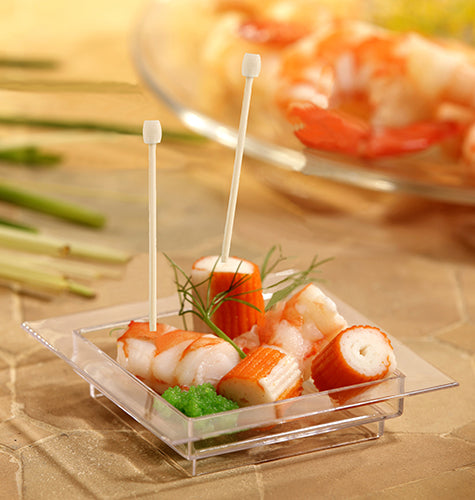 Eine kleine quadratische durchsichtige Schale enthält Krabbenimitat, grüne Garnierung und zwei PAPSTAR 100 Fingerfood - Picker von PAPSTAR GmbH. Unscharfes Gemüse und weitere Meeresfrüchte erscheinen im Hintergrund.