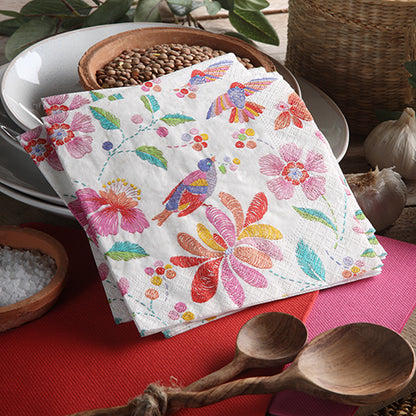 A stack of PAPSTAR 20 napkins, 25 x 25 cm, 3-ply, 1/4-fold from PAPSTAR GmbH with colorful flower and bird motifs lies on a table between wooden spoons, grains, and a basket – perfect as a vibrant table decoration. Plates in the background.