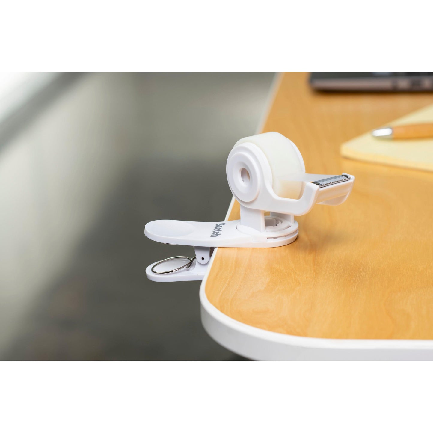 Der Scotch™ Clip & Twist Klebeband-Abroller C19 mit Scotch® Magic™ Unsichtbares Klebeband (19 mm × 8,89 m) von 3M Deutschland GmbH wird neben einem Notizblock und einem Stift an einen Holztisch geklemmt.