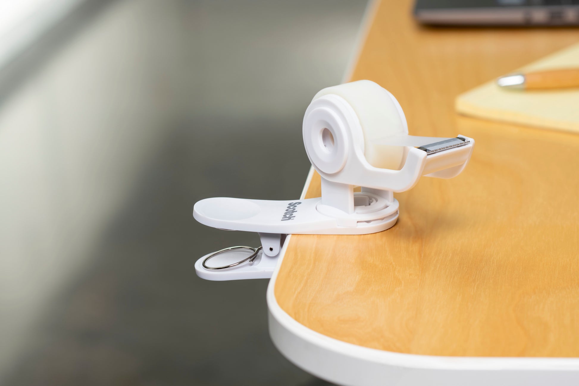 Der Scotch™ Clip & Twist Klebeband-Abroller C19 mit Scotch® Magic™ Unsichtbares Klebeband (19 mm × 8,89 m) von 3M Deutschland GmbH wird neben einem Notizblock und einem Stift an einen Holztisch geklemmt.