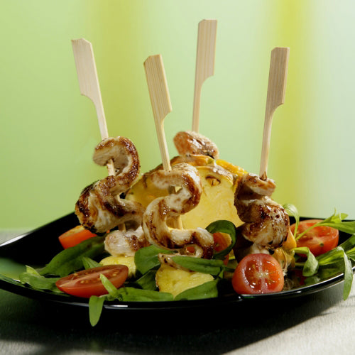 Ein schwarzer Teller mit gegrillten Hähnchenspießen auf Grünzeug und Kirschtomaten, arrangiert mit PAPSTAR 250 Fingerfood - Spieße "pure" von PAPSTAR GmbH rund um die Ananas - perfekt für jedes Partybuffet. Drei Holzspieße werden in die Ananas gesteckt.