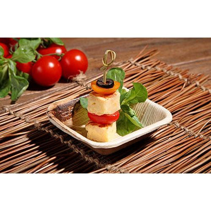 On a small bamboo plate lies a skewer with cheese, tomatoes, peppers, olives, and vegetables. PAPSTAR 25 bowls, palm leaf "pure" round from PAPSTAR GmbH are perfectly suited for sustainable events due to their eco-friendly design.