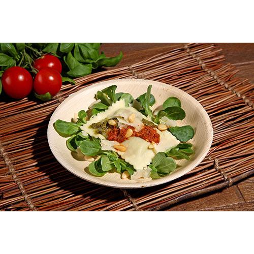 Ein Teller Ravioli mit Tomatensoße, Pinienkernen und Käse auf Blattgemüse wird auf einem PAPSTAR 25 Teller Palmblatt „pure“ rund der PAPSTAR GmbH serviert, der auf einem Bambus-Tischset mit Kirschtomaten und Blattgemüse im Hintergrund ruht.