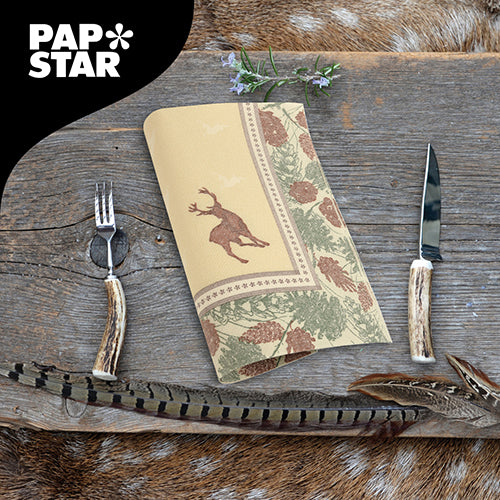 Eine Serviette der PAPSTAR GmbH ROYAL Collection (40x40 cm, 1/4-faltig) mit Hirsch- und Tannenzapfenmotiv liegt auf einem rustikalen Holztisch mit geweihähnlichem Besteck, Federn und Kräutern. In der linken oberen Ecke befindet sich das PAPSTAR-Logo.