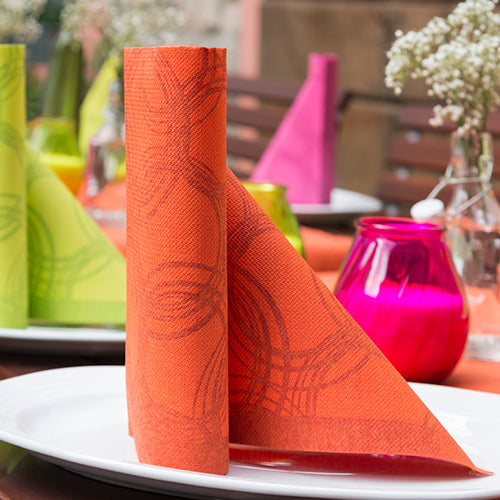 Eine ordentlich gefaltete orangefarbene PAPSTAR 50 Servietten "ROYAL Collection" 1/4-Falz 40 x 40 cm nektarine Serviette steht aufrecht auf einem weißen Teller, mit buntem ROYAL Collection-Dekor und Blumen im Hintergrund verschwommen.