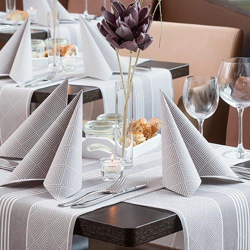 Ein mit PAPSTAR GmbH 50 Servietten "ROYAL Collection" 1/4-Falz, 40 x 40 cm, braun, in Dreiecken gefaltet, gedeckter Restauranttisch mit Weingläsern, Besteck, einem Brotkorb, einem violetten Kunstblumen-Mittelstück und einem brennenden Teelicht.