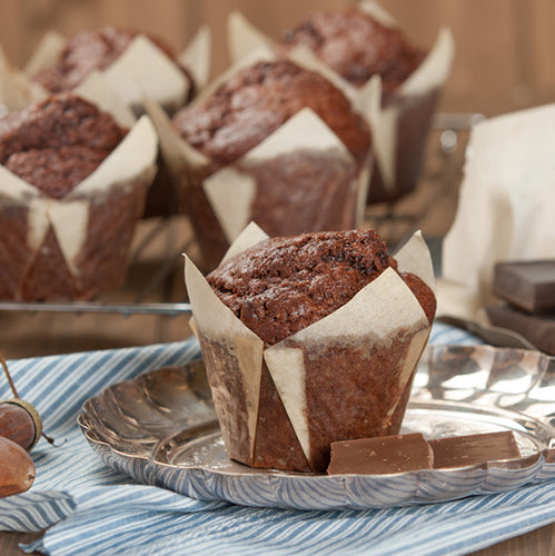 Ein Schokomuffin im weißen PAPSTAR 200 Backförmchen „Tulip“ der PAPSTAR GmbH liegt neben zwei Pralinen auf einem silbernen Teller. Im Hintergrund sind weitere Muffins, Cupcakes und ein Kuchengitter zu sehen, unter dem Teller befindet sich ein blau gestreiftes Tuch.