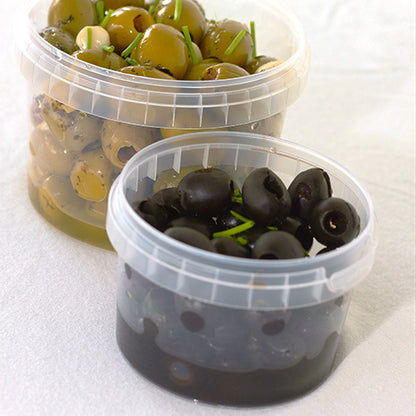On a white background, there are two transparent Starpak 40 delicatessen cups (PP, 480 ml, Ø11.8cm, with shrink wrap and tamper-evident closure) - one with green olives in brine and herbs, the other with sliced black olives, both garnished with chives.