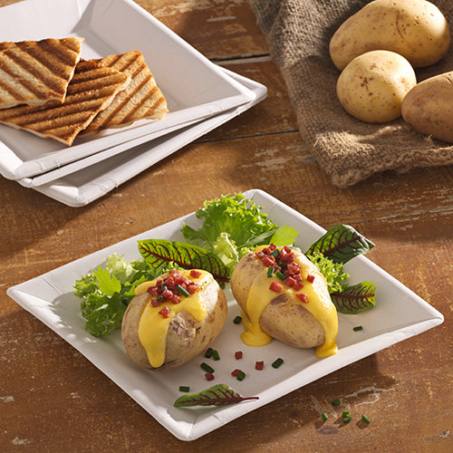 Zwei Ofenkartoffeln mit gelber Käsesauce, Speckwürfeln und Schnittlauch werden auf PAPSTAR-Papptellern (22,5 x 22,5 cm) (PAPSTAR GmbH) mit Blattgemüse serviert. Im Hintergrund stehen gegrilltes Brot und ganze Kartoffeln auf einem Holztisch.