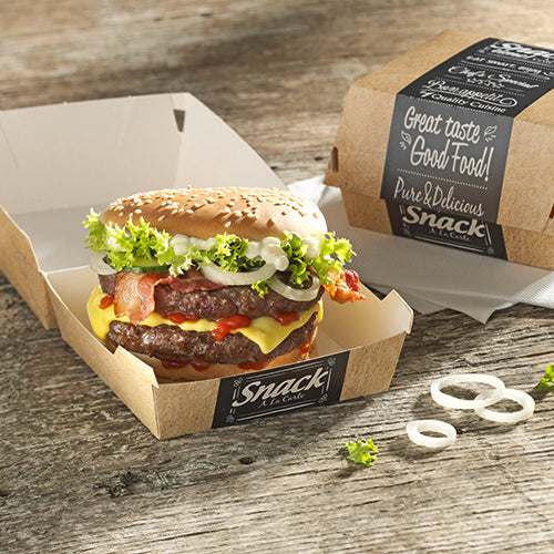 Ein Cheeseburger mit Salat, Tomate, Bacon, Zwiebeln und Sauce liegt in einer offenen PAPSTAR 50 Burgerbox aus Pappe "pure" von PAPSTAR GmbH auf einem Holztisch. Daneben eine geschlossene Box mit "Good Food"-Aufdruck und Onion Rings.