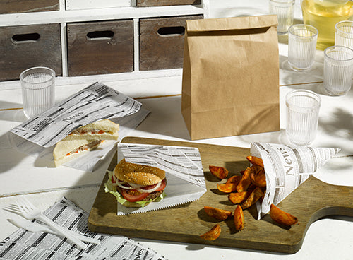 Ein Sandwich, ein Burger in PAPSTAR Hamburger-Tüten Pergament-Ersatz "Newsprint" (16x18 cm, 300 Stück) und Kartoffelecken liegen auf einem Holzbrett. Daneben stehen durchsichtige Becher, weißes Besteck und eine braune Papiertüte auf dem Tisch. Marke: PAPSTAR GmbH.
