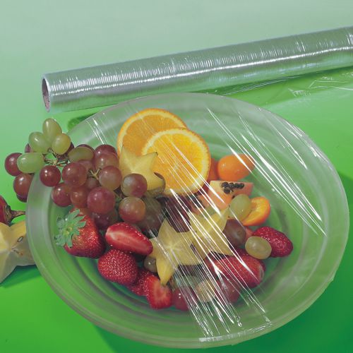 A transparent Starpak cling film folding box covers a plate with fresh grapes, strawberries, orange slices, and star fruits to keep the food fresh. A roll of Starpak cling film can be seen on a green surface in the background.