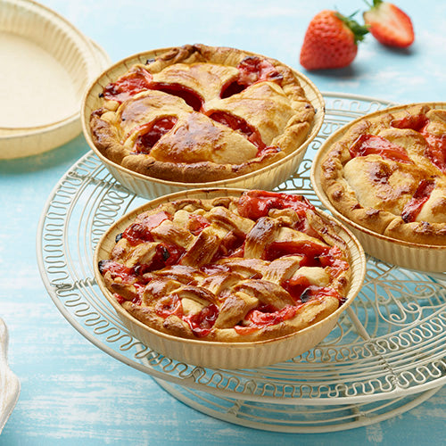 Drei kleine Erdbeertörtchen mit goldener Kruste, teilweise mit Gitteraufsätzen, sitzen auf einem weißen Drahtkuchenständer. Im Hintergrund sind zwei frische Erdbeeren und eine PAPSTAR 25 Backformen "pure" rund braun von PAPSTAR GmbH auf hellblauem Untergrund zu sehen.