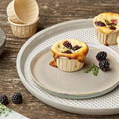 A blackberry muffin in a PAPSTAR 50 baking molds "pure" brown from PAPSTAR GmbH lies on a gray plate, with more muffins, blackberries, and thyme on a wooden table. In the background, stacked empty baking molds can be seen.