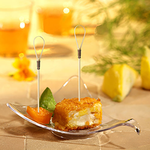 Auf einem kleinen, durchsichtigen, blattförmigen Schälchen liegen ein panierter, angebissener Vorspeisenwürfel, ein PAPSTAR 40 Fingerfood - Picker 10 cm „Loop“ der PAPSTAR GmbH, ein orangefarbenes Fruchtstück und grüne Garnitur. Im Hintergrund verschwimmen Getränke und Zitronenspalten.