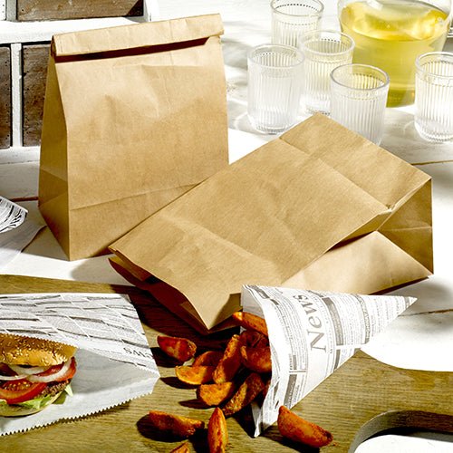 Two PAPSTAR 250 block bottom bags made of brown kraft paper from PAPSTAR GmbH, a burger in newspaper, and a portion of potato wedges on wood. In the background are four glasses and a jug.