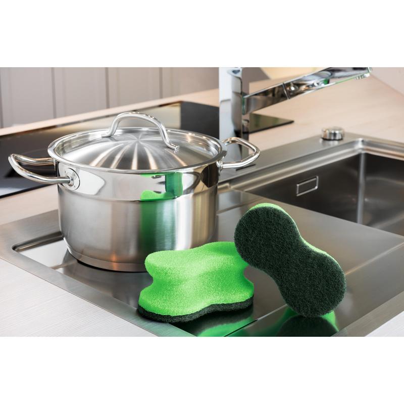 A stainless steel pot with a lid stands on the kitchen counter next to the sink. Two Scotch-Brite® Classic Cleaning Sponges from 3M Deutschland GmbH are placed in the foreground next to the pot.