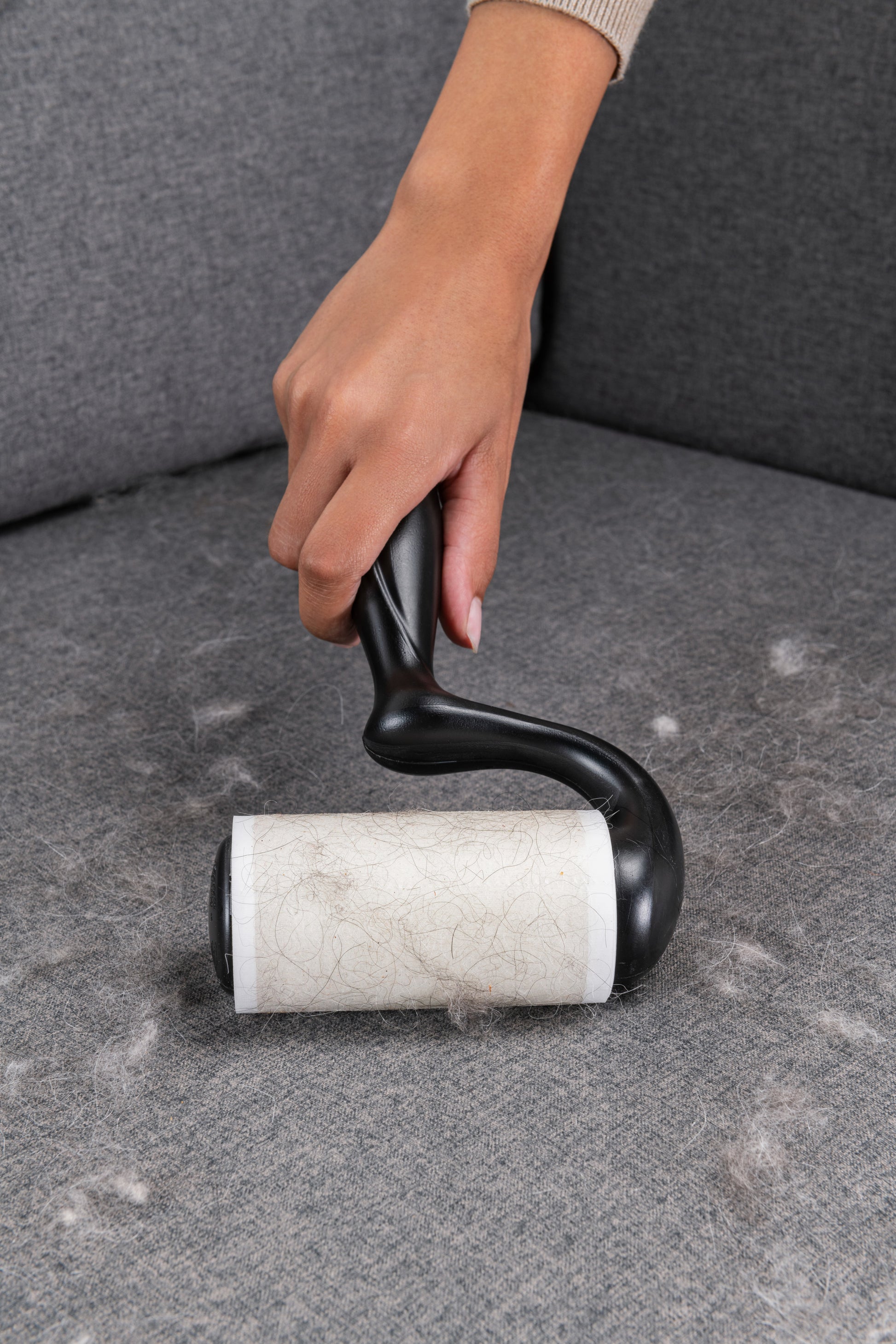 Eine Hand verwendet den Scotch-Brite™ Everyday Clean Fusselroller für flache Oberflächen (56 Blatt, 1 Packung) von 3M Deutschland GmbH, um Tierhaare von einer grau gepolsterten Couch zu entfernen. Der Roller ist teilweise mit hellen Haaren bedeckt.