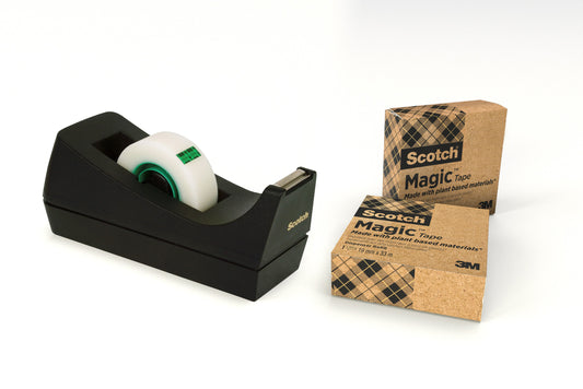 Ein schwarzer Scotch® C38 Klebebandabroller mit einer Rolle Magic™ Klebeband von 3M Deutschland GmbH steht neben zwei Kartons mit der Aufschrift "Scotch® Magic™", jede Rolle mit 66 % pflanzlichem Klebstoff, Größe 19 mm x 33 m.