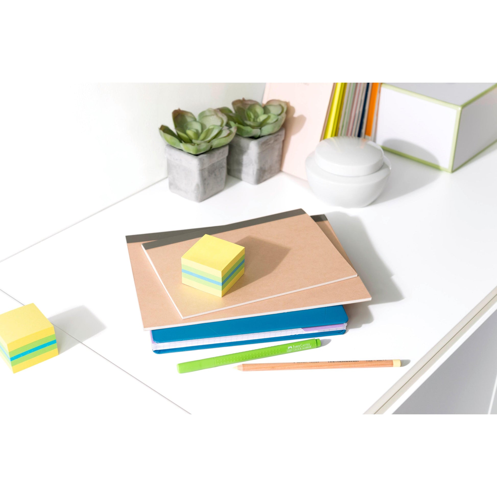 Auf einem weißen Schreibtisch stehen zwei getopfte Sukkulenten, Notizbücher, ein grüner Stift, ein Bleistift, ein weißer Behälter, bunte Mappen und Schachteln. Auf dem Schreibtisch liegen 3M Deutschland GmbH Post-it® Notes Mini Würfel (51 mm x 51 mm, 400 Blatt/Block, 100% PEFC).