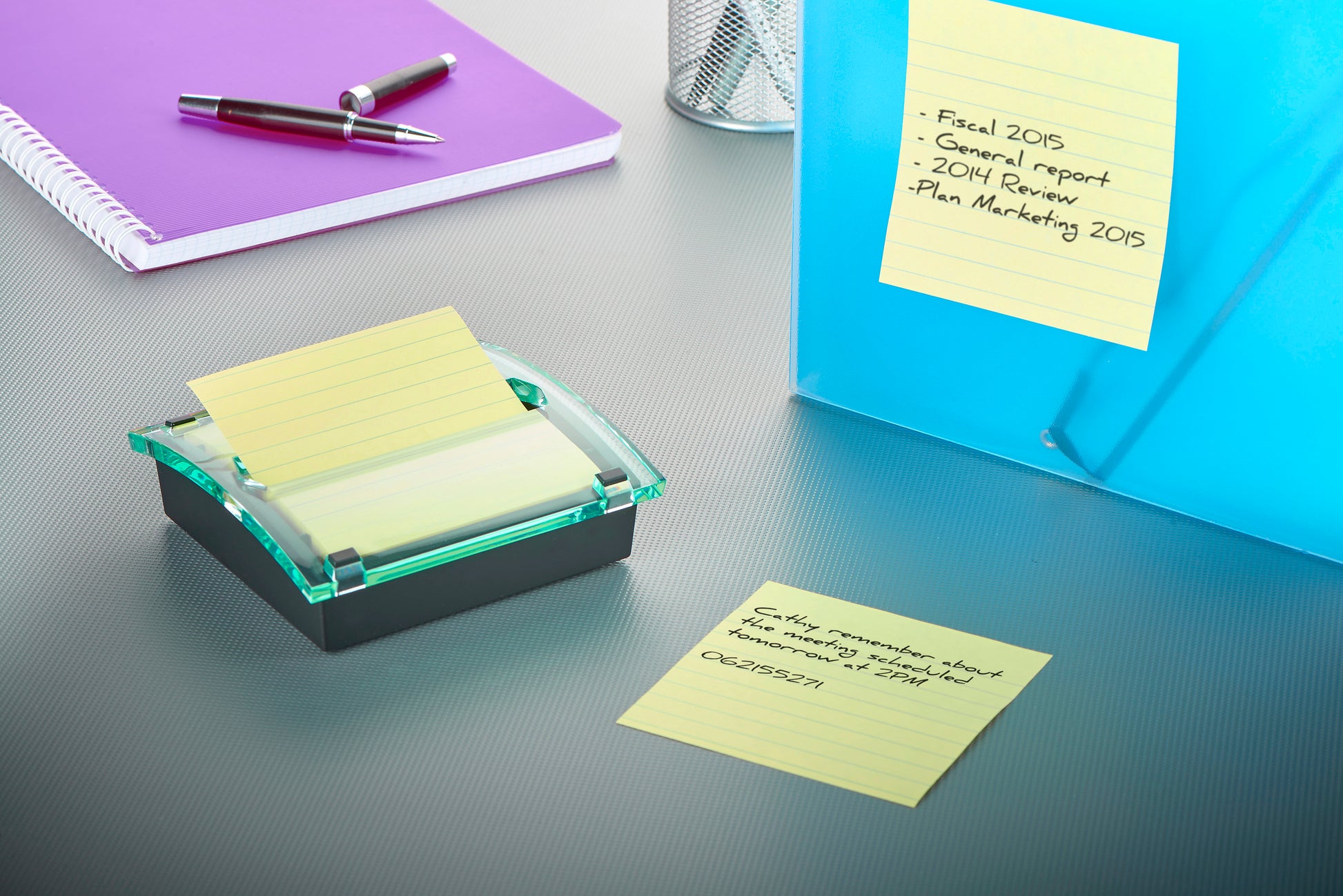 Auf einem grauen Schreibtisch liegen ein lila Notizbuch, ein silberner Stift, ein 3M Post-it® Z-Notes Dispenser XL (schwarz) mit Super Sticky Z-Notes (gelb, liniert, 101x101mm), handgeschriebene Haftnotizen, eine blaue Mappe und ein Metallnetz-Stifthalter.