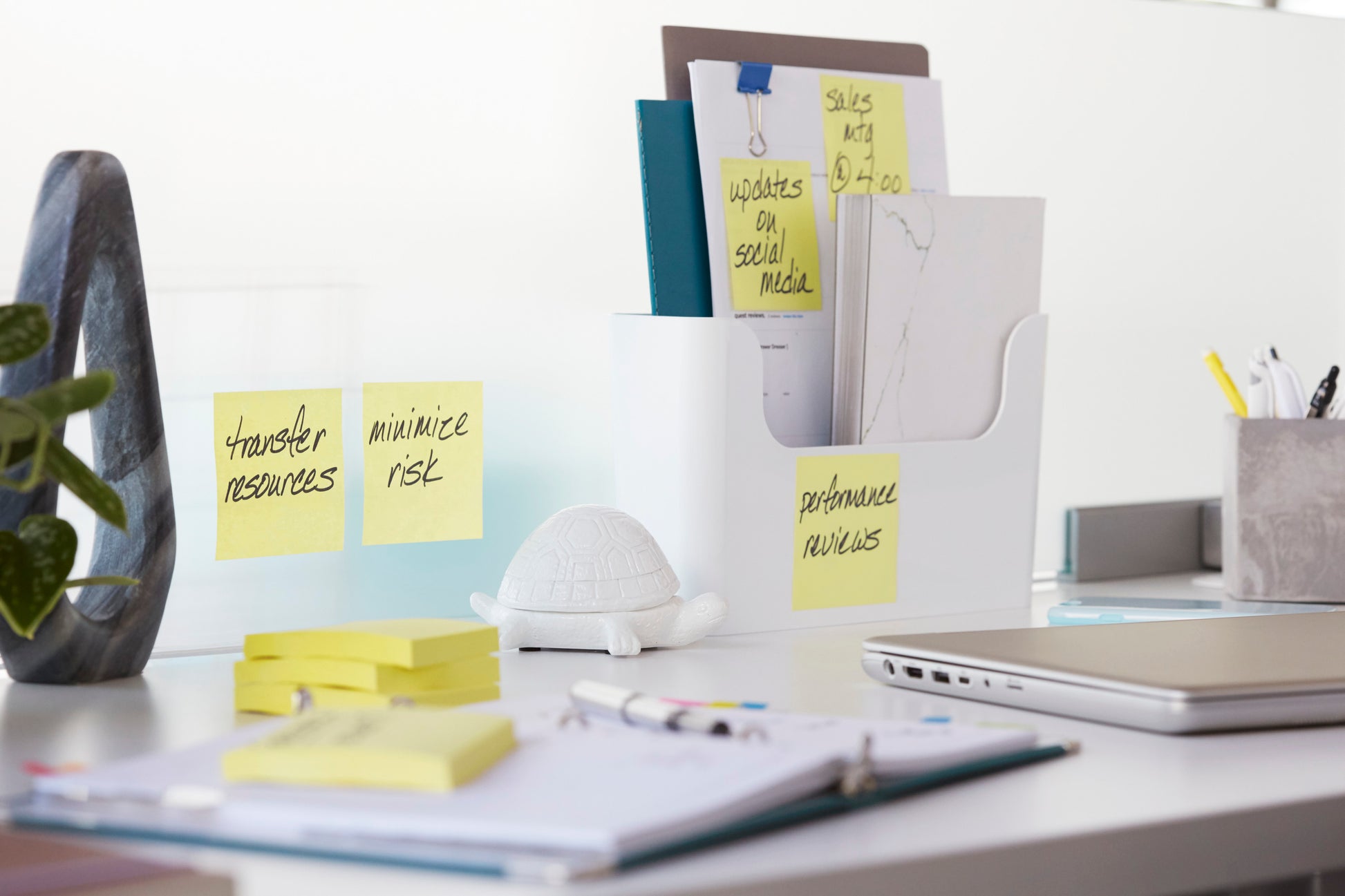 Ein weißer Schreibtisch mit dem Post-it® Z-Notes Dispenser PRO der 3M Deutschland GmbH in Schwarz mit gelben Post-it® Super Sticky Z-Notes (76x76 mm), dazu Mappen, Büromaterial, ein Notebook, ein Laptop, eine Schildkrötenfigur und Haftnotizen an der Wand, auf denen Aufgaben aufgelistet sind.