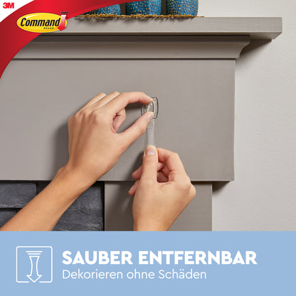 Eine Person zieht einen transparenten Klebestreifen von einem grauen Kaminsims ab und zeigt, wie Command™ Mini-Haken, transparent, mit transparenten Strips (17006CLR) von 3M Deutschland GmbH spurlos dekorieren und befestigen. Command und 3M Logos sind sichtbar.