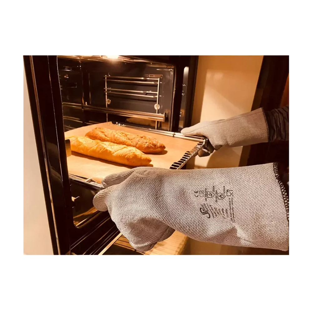 A person with AMPri SolidSafety Heat Protectors heat protection gloves L - XXL from AMPri Handelsgesellschaft mbH carefully removes a baking tray from the oven. On the tray are two loaves of bread, while the warm lighting of the kitchen highlights both the open oven and the maximum heat protection of these protective gloves.