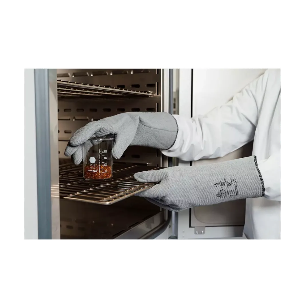 A person wears AMPri SolidSafety heat protection gloves L - XXL from AMPri Handelsgesellschaft mbH and a lab coat and carefully places a glass beaker with reddish liquid on a metal stand in an industrial oven. The focus is on the hands and the beaker, emphasizing the maximum heat protection, with the shelves inside the device visible.