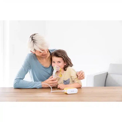 An adult helps a child use the discontinued item: Beurer small inhaler IH 58 KIDS from Beurer GmbH at a wooden table. The child holds the mask to their face, both sitting in a bright, minimalist room.