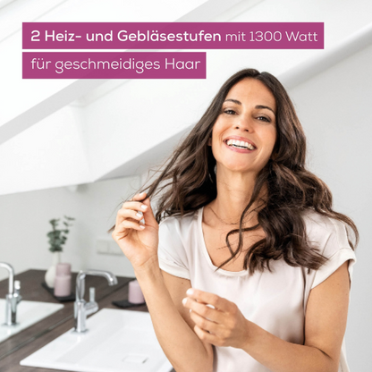 A woman with long, wavy hair smiles while holding a strand of hair. The background shows a modern bathroom with a sink and a potted plant. The text on the image reads: “Beurer HC 17 Foldable Compact Hairdryer by Beurer GmbH: 2 heat and blower settings with 1300 watts for smooth hair”, which means “Powerful compact hairdryer”.