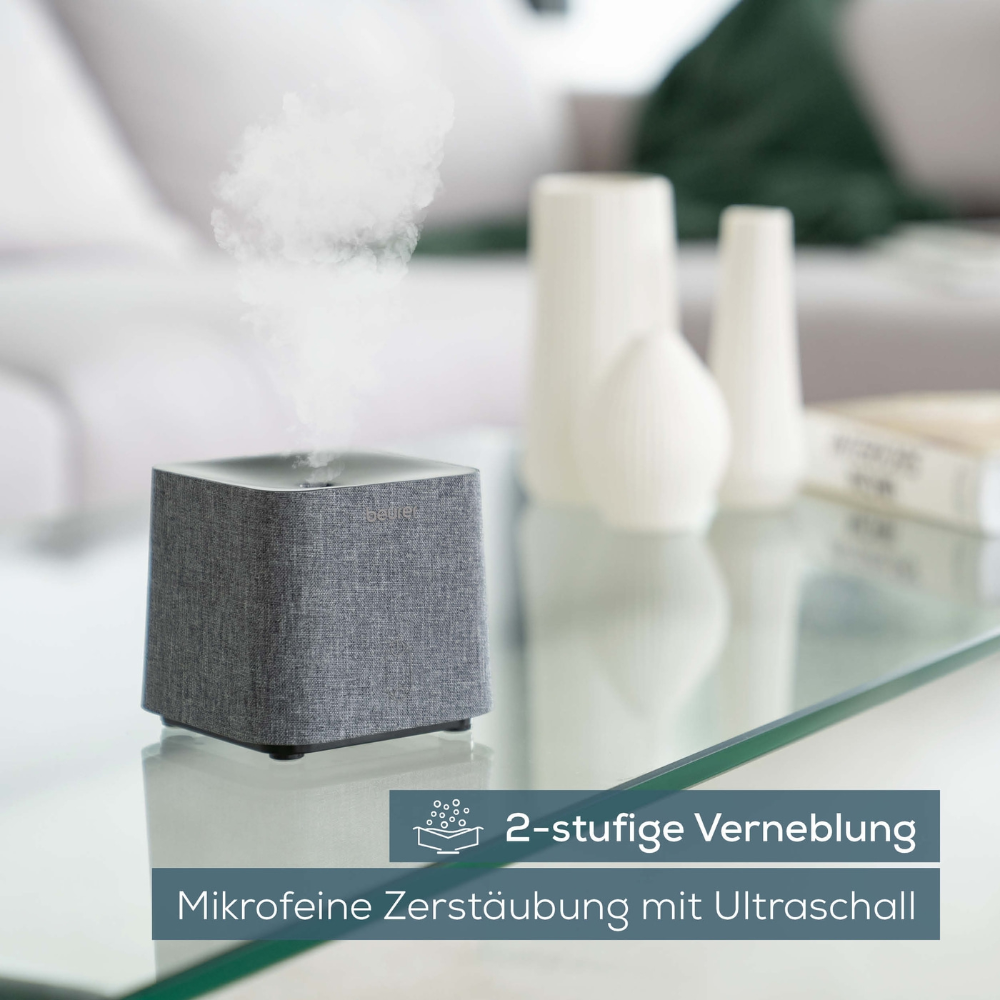 Ein Beurer LA 35 Aroma-Diffusor der Beurer GmbH, frei in Grau, schmückt einen Glastisch in einem mit Möbeln geschmückten Wohnzimmer. Der Text auf dem Bild lautet „2-stufige Verneblung“ und „Mikrofeine Zerstäubung mit Ultraschall“, was die zweistufige Vernebelung und ultrafeine Zerstäubung mittels Ultraschalltechnologie hervorhebt. Dieses vielseitige Gerät fungiert sowohl als Luftbefeuchter als auch als Aroma-Diffusor.