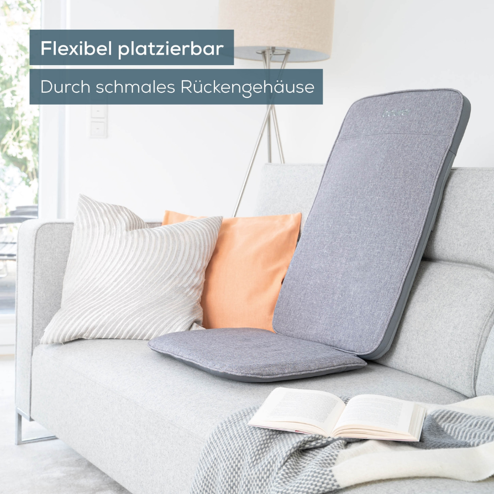 Eine graue Beurer MG 202 Shiatsu-Massagesitzauflage der Beurer GmbH steht auf einem hellen Sofa, das mit orangefarbenen und weißen Kissen geschmückt ist. Auf dem Sofa liegen ein aufgeschlagenes Buch und eine Decke, im Hintergrund steht eine Stehlampe. Der Text lautet: „Flexibel platzierbar durch schmales Rückengehäuse.“