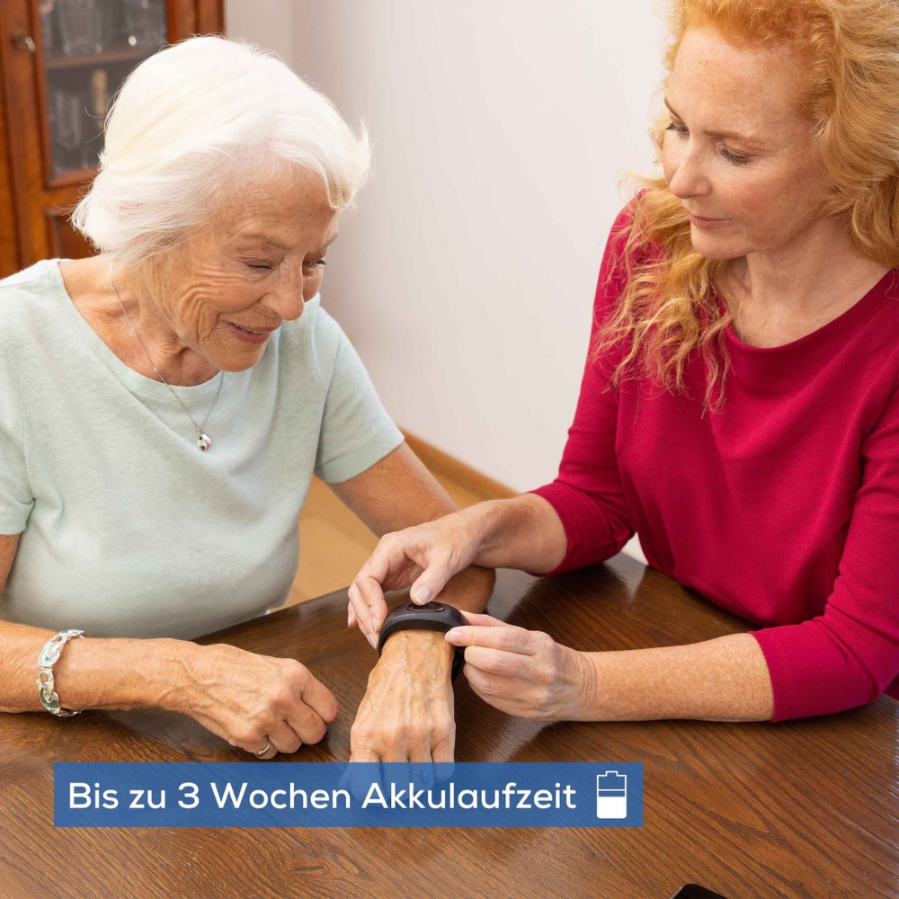 Zwei Frauen sitzen an einem Holztisch. Eine Frau in einem rosa Hemd demonstriert einer älteren Frau in einem hellblauen Hemd ein Beurer Notfallarmband EC 70 SafePlus | Packung (1 Stück) von Beurer GmbH. Der Text auf dem Bild lautet auf Deutsch „Bis zu 3 Wochen Akkulaufzeit“, was auf eine Akkulaufzeit von bis zu drei Wochen hinweist.