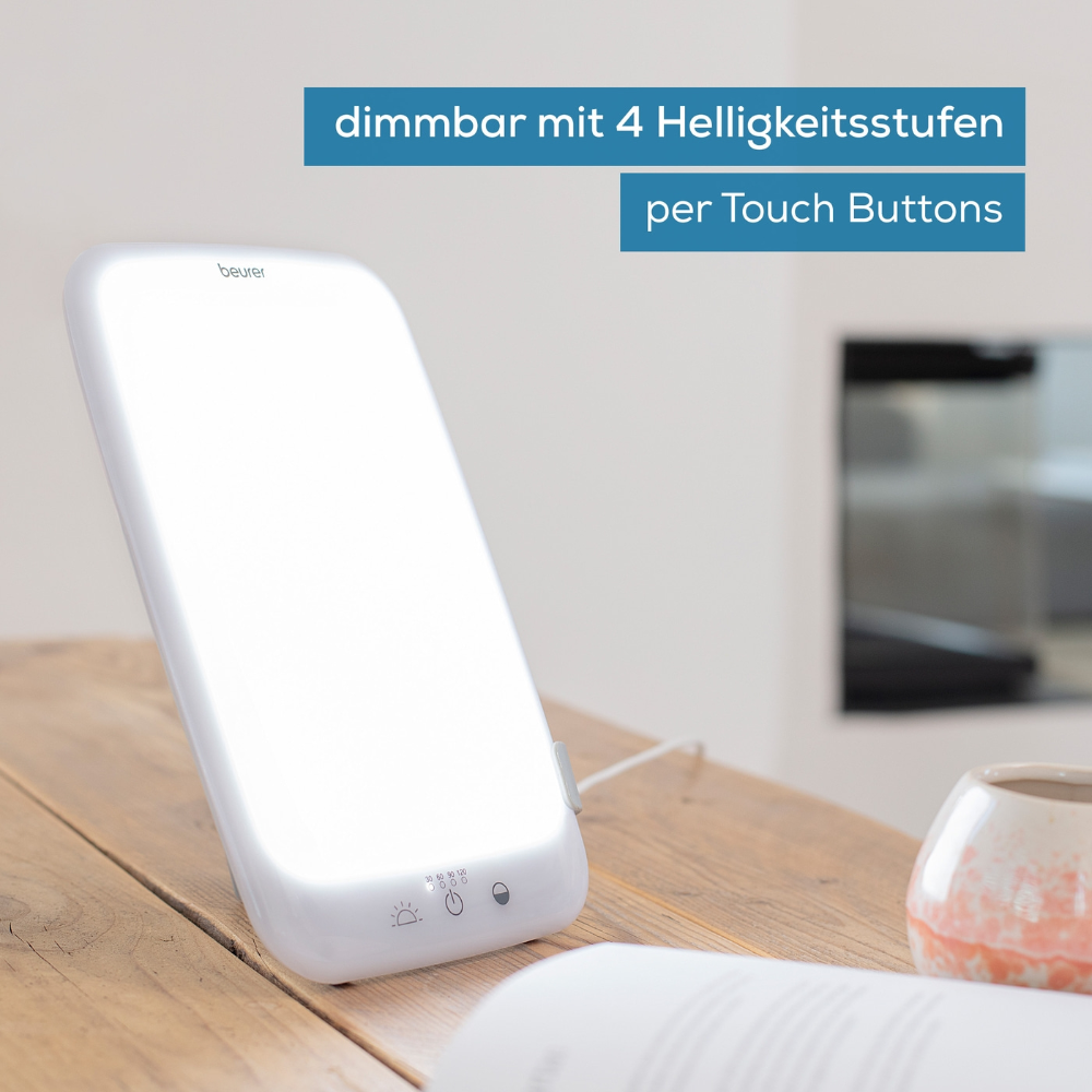 Eine rechteckige Beurer Tageslichtlampe TL 35 der Beurer GmbH steht auf einem Tisch. Der Text auf dem Bild lautet „dimmbar mit 4 Helligkeitsstufen per Touch Buttons“. Im Hintergrund ist eine verschwommene Wohnzimmerkulisse zu sehen. Die Lichttherapielampe ist beleuchtet und an eine Stromquelle angeschlossen.